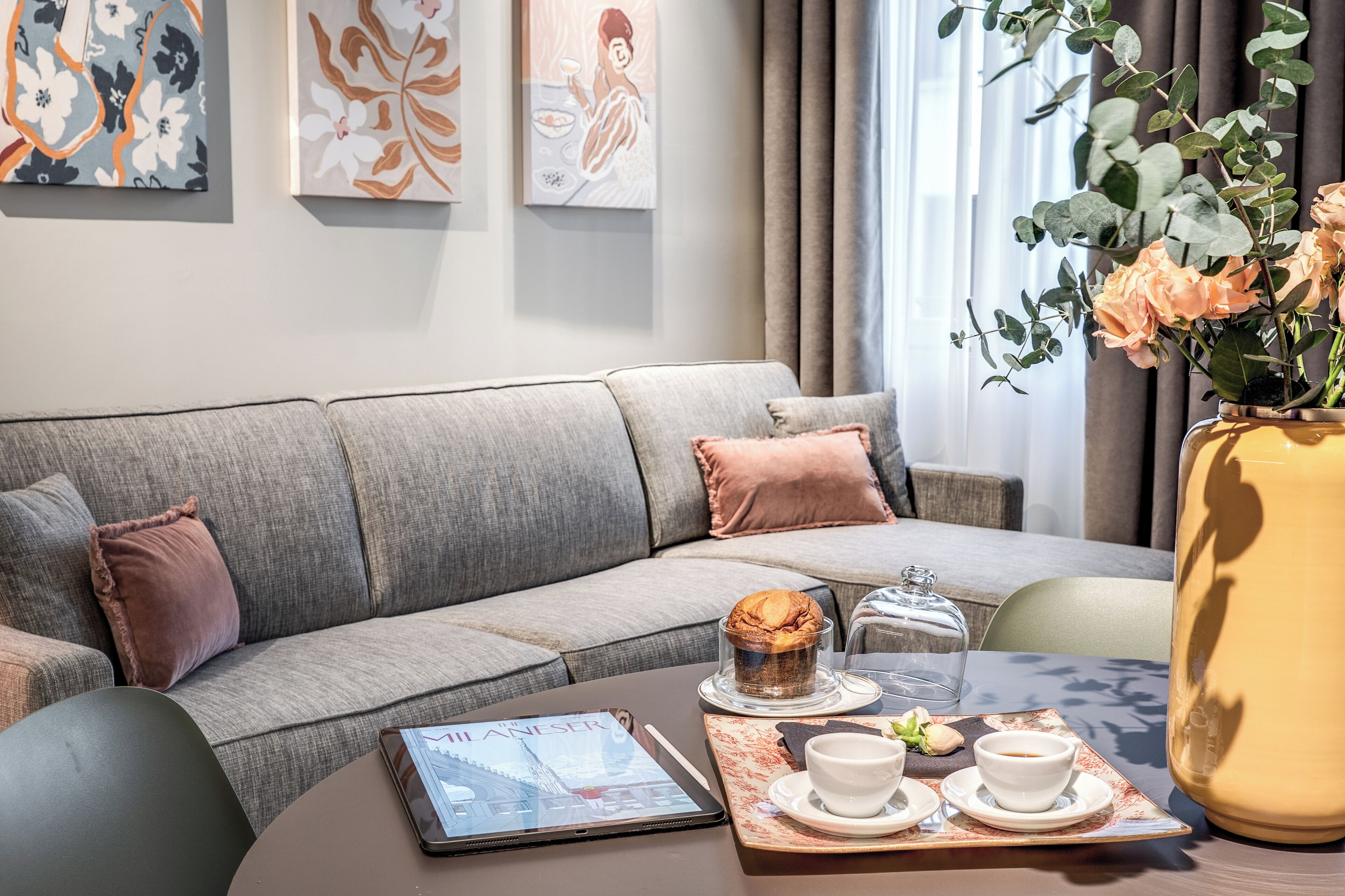 Una sala de estar moderna con un sofá gris, una mesa de centro con un muffin, una tableta y una bandeja con tazas y galletas.
