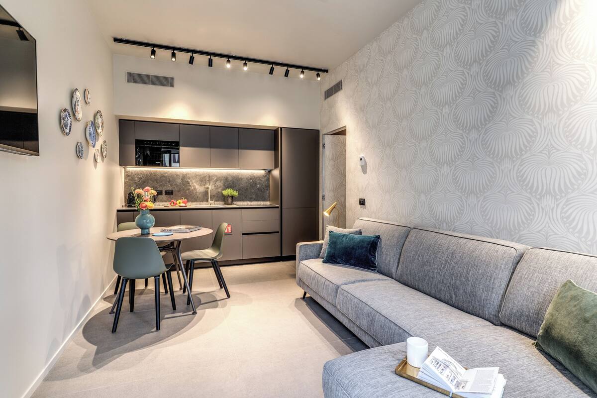 A modern living room with a grey sofa, a dining table with chairs, and a kitchen area with dark cabinets and a marble countertop.