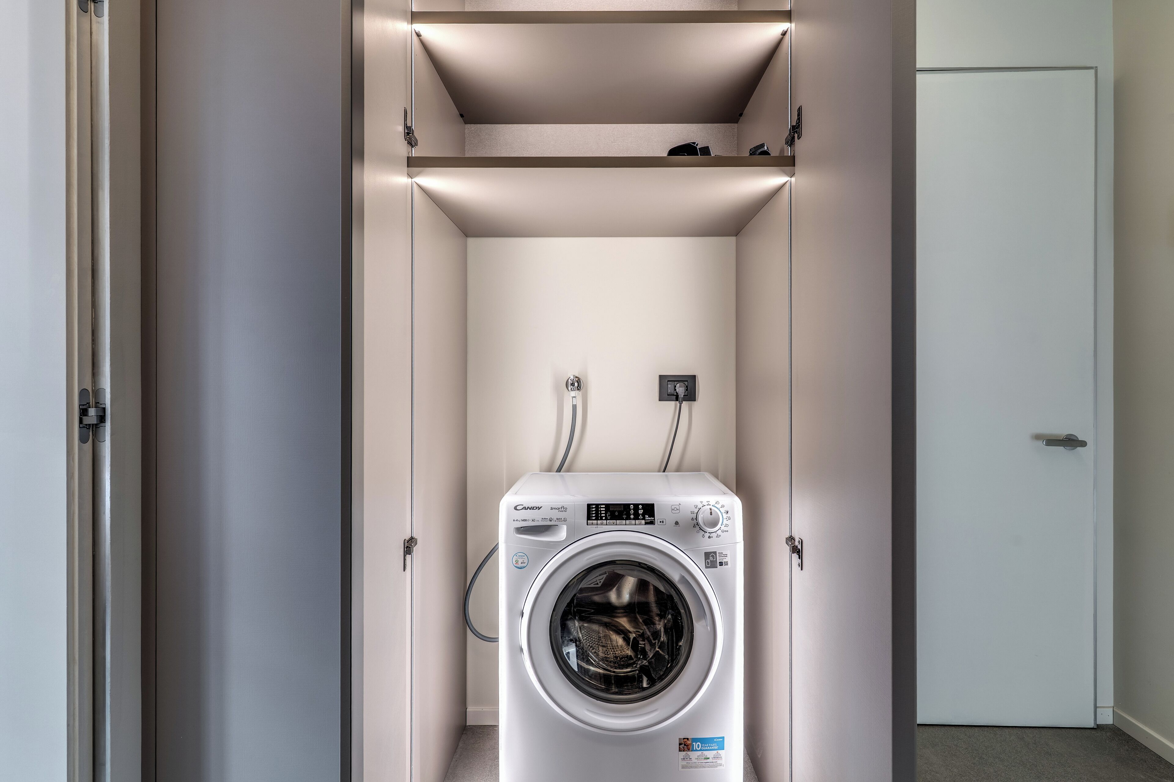 A compact laundry room with a front-loading washing machine, a small shelf above, and a door to the right.