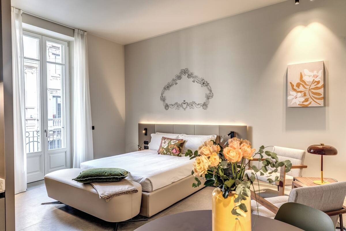 A modern hotel room with a bed, a sofa, a dining table, and a vase of flowers.