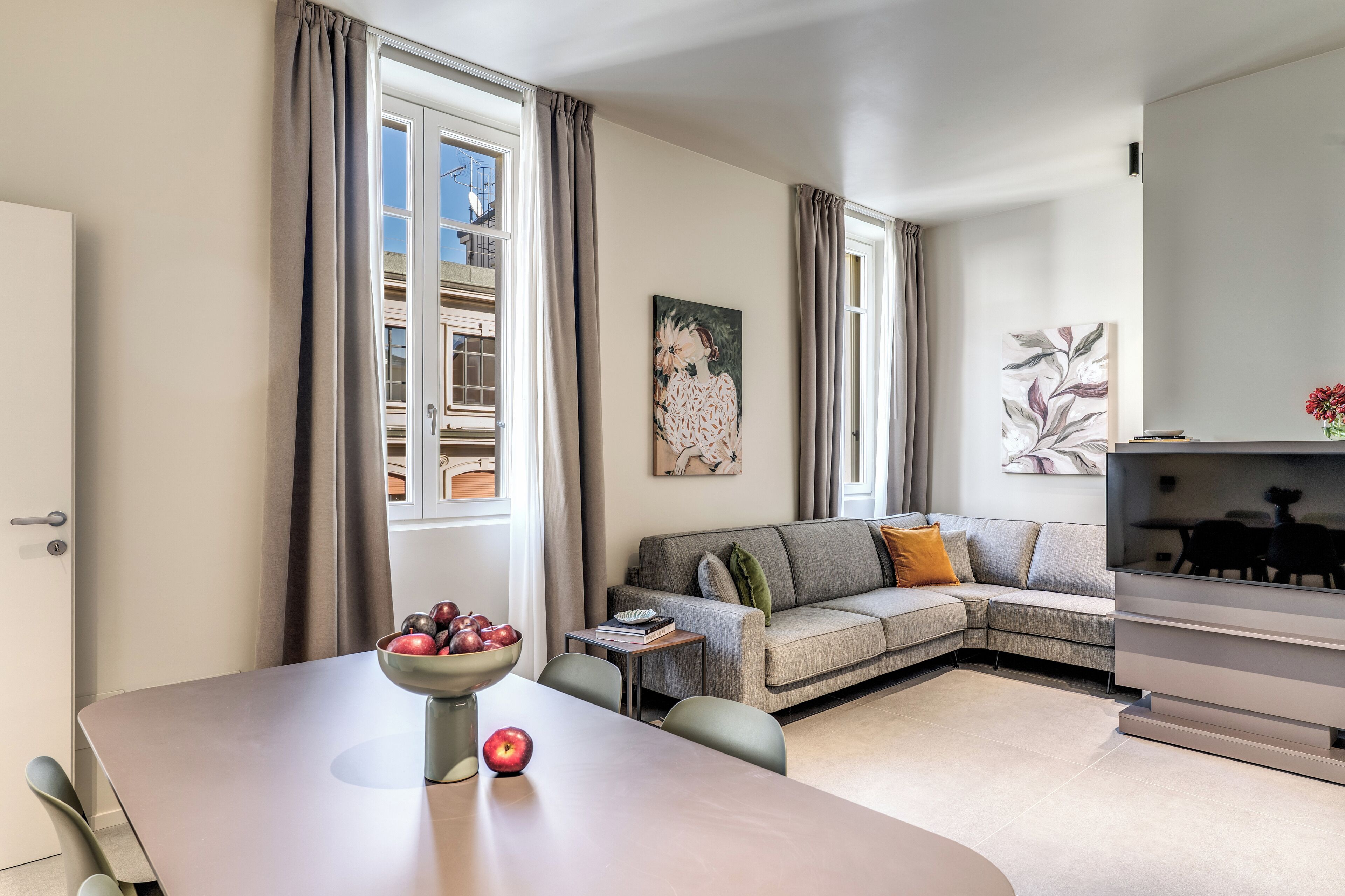 A modern living room with a sofa, a dining table with a bowl of fruit, and a flat-screen TV.