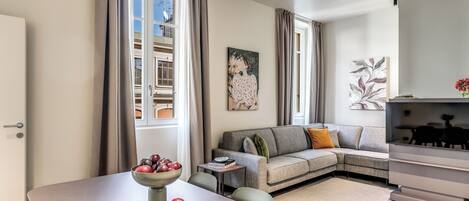 A modern living room with a sofa, a dining table with a bowl of fruit, and a flat-screen TV.