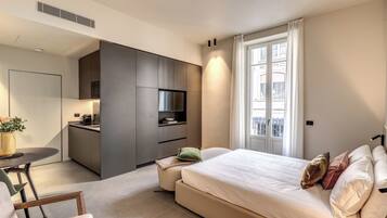 A modern hotel room with a bed, a small kitchenette, a dining area, and a large window with curtains.