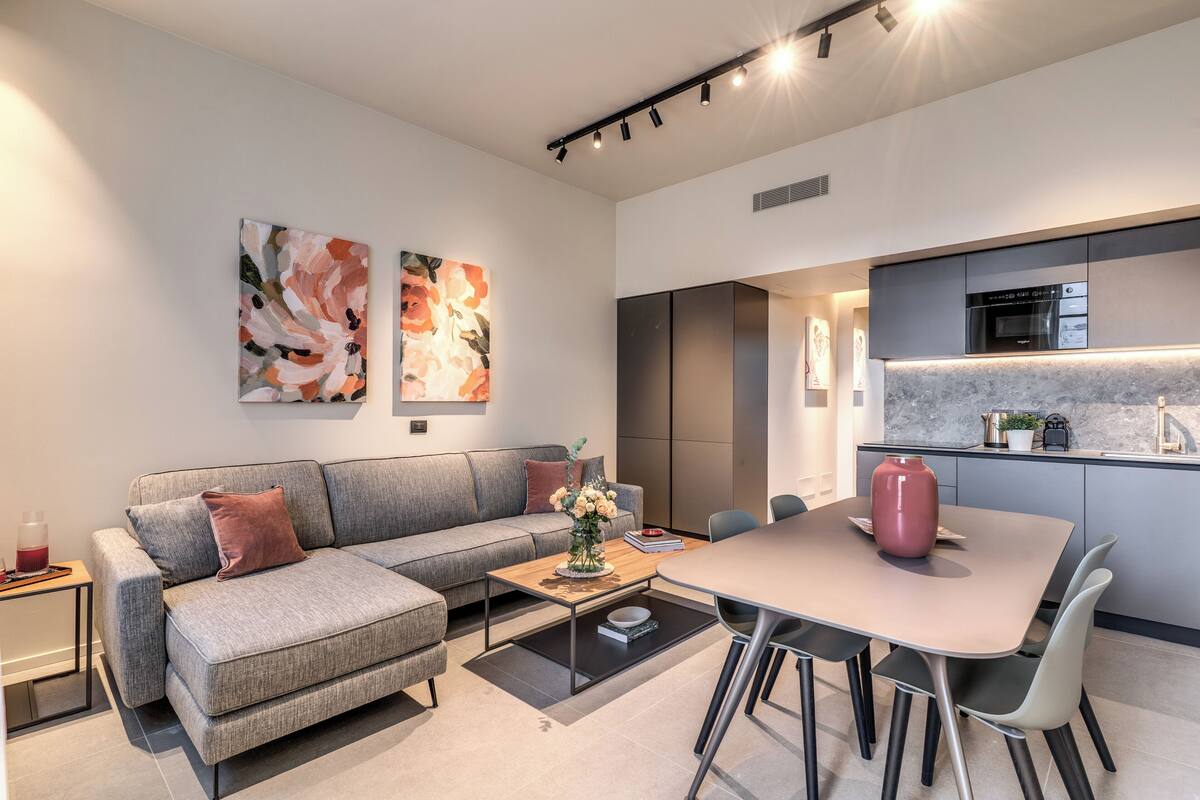 A modern living room with a grey sofa, a coffee table, and a dining area with a table and chairs. There is a kitchen area with a stove and a sink.