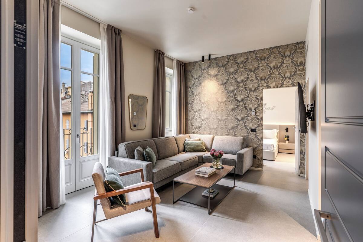 A modern living room with a sofa, coffee table, and a chair. There is a patterned wallpaper on the wall and a window with curtains.