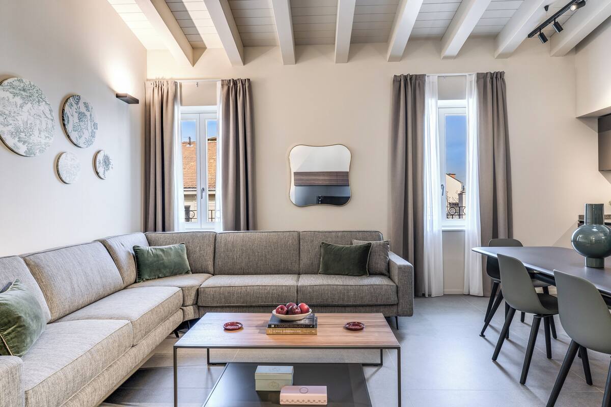 A modern living room with a sectional sofa, a coffee table with books and a bowl of fruit, and a dining area with a table and chairs.
