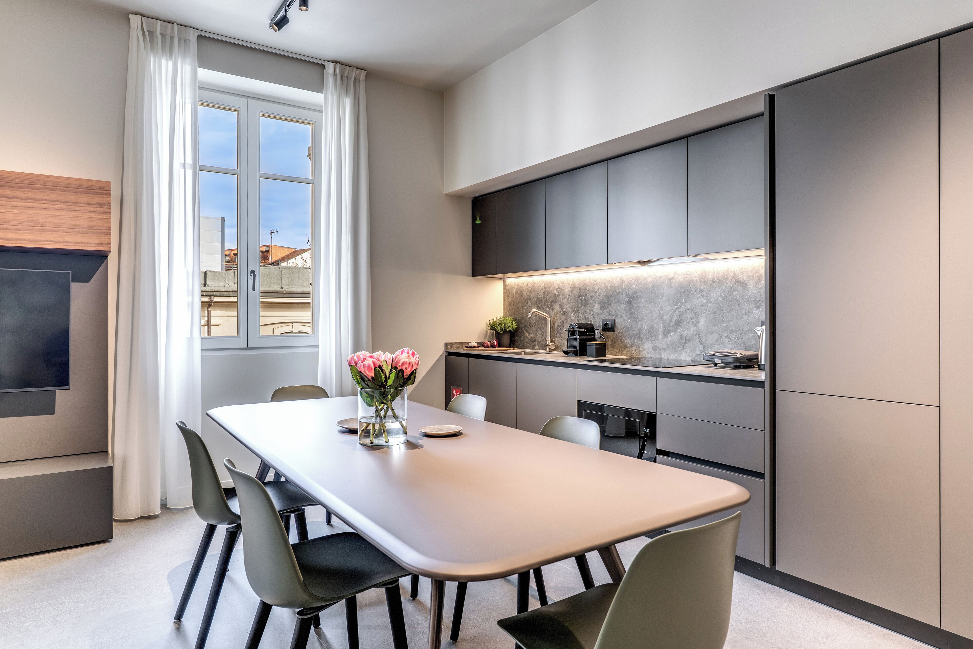 A modern kitchen with a dining area, featuring a large table, chairs, and a vase of flowers.