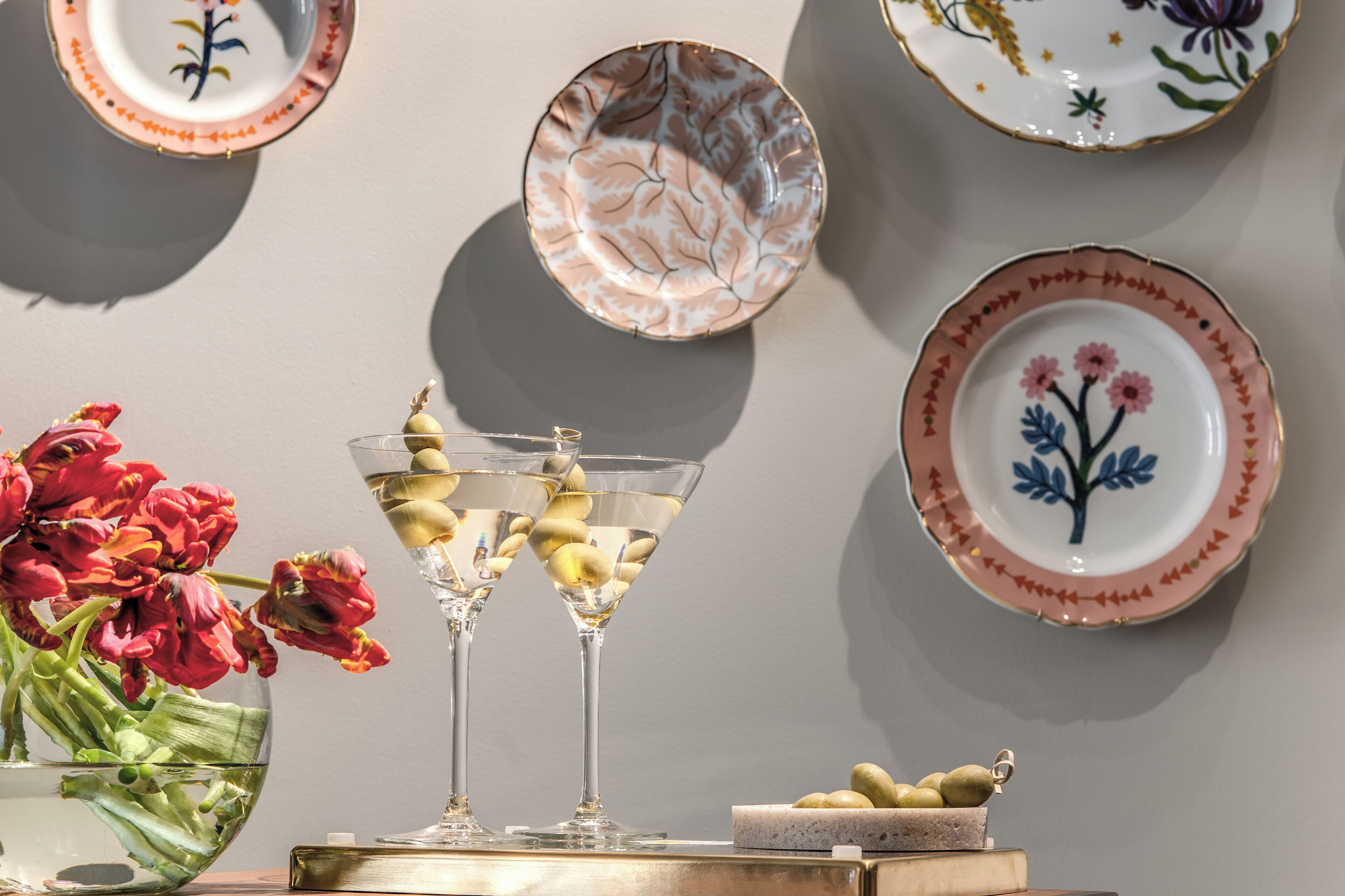 Una mesa con dos copas de martini, un jarrón de flores rojas y un plato de aceitunas.