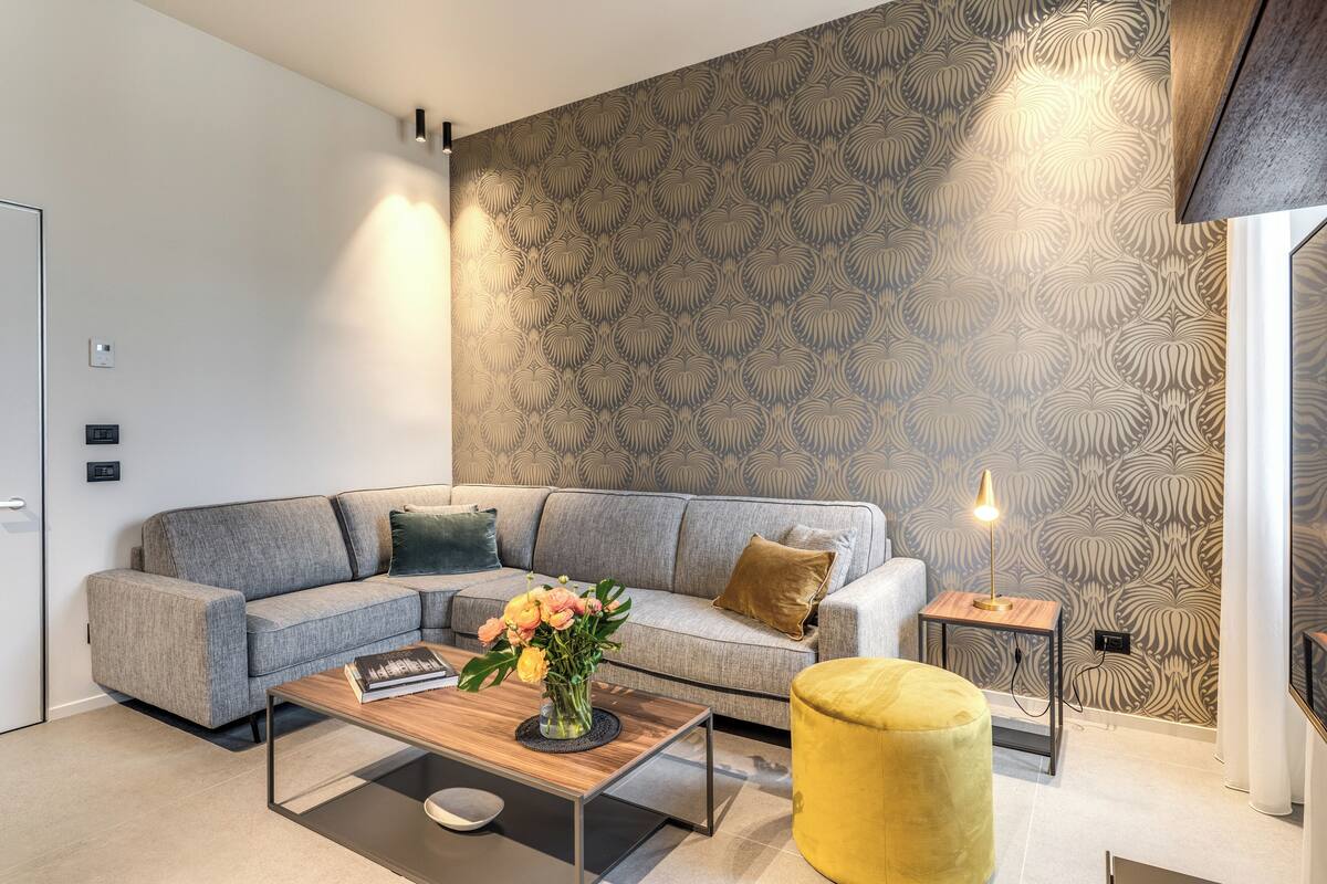 A modern living room with a grey sofa, a wooden coffee table, a yellow ottoman, and a patterned wallpaper.