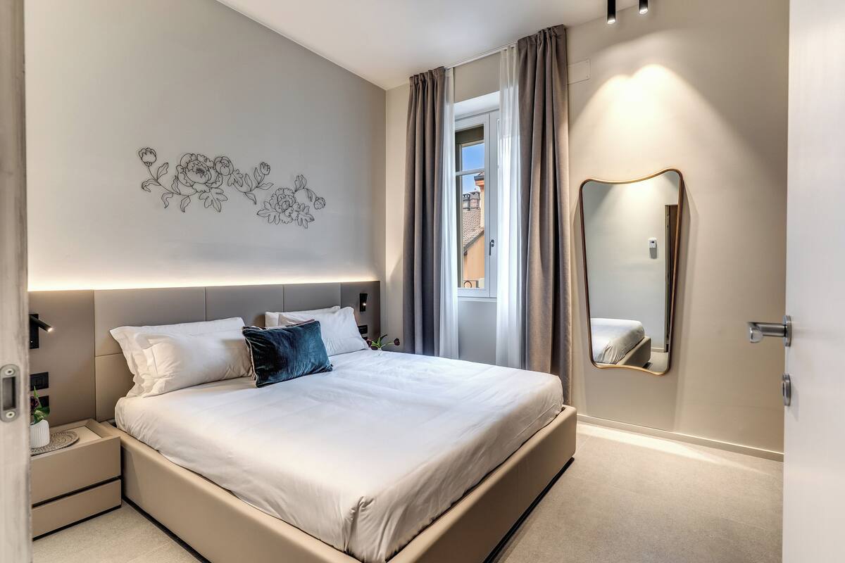 A modern bedroom with a large bed, a nightstand, a wall-mounted mirror, and a window with curtains.