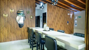 Un comedor moderno con una larga mesa blanca, sillas negras, paneles de madera y lámparas colgantes geométricas.