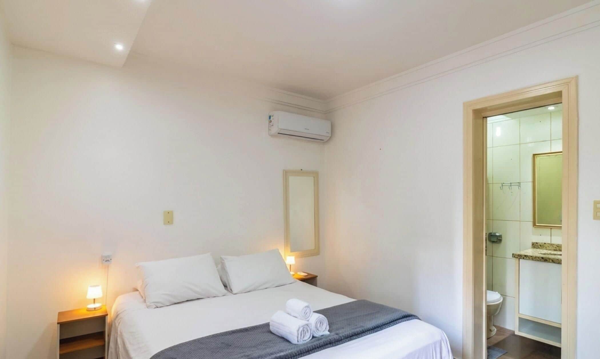 A hotel room with a bed, bedside lamps, an air conditioning unit, a mirror, and a bathroom with a toilet and sink.