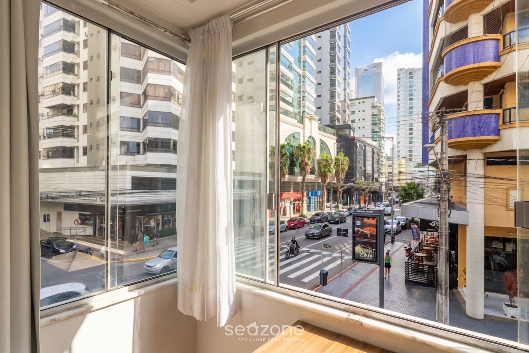 A room with a large window overlooking a busy street, a bed, and a view of tall buildings.