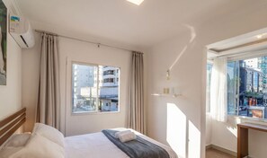 Une chambre d’hôtel avec un lit, une fenêtre avec des rideaux, un climatiseur fixé au mur et une vue sur les bâtiments environnants.