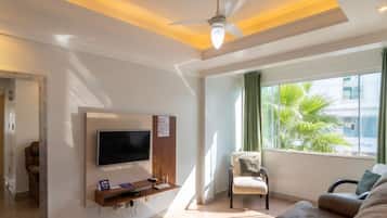 Una sala de estar con televisor de pantalla plana, ventilador de techo, ventana con vista a palmeras y una zona de estar con sillón y sofá.