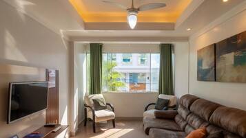 Una sala de estar con televisor de pantalla plana, ventilador de techo, sofá, sillón y ventana con cortinas.