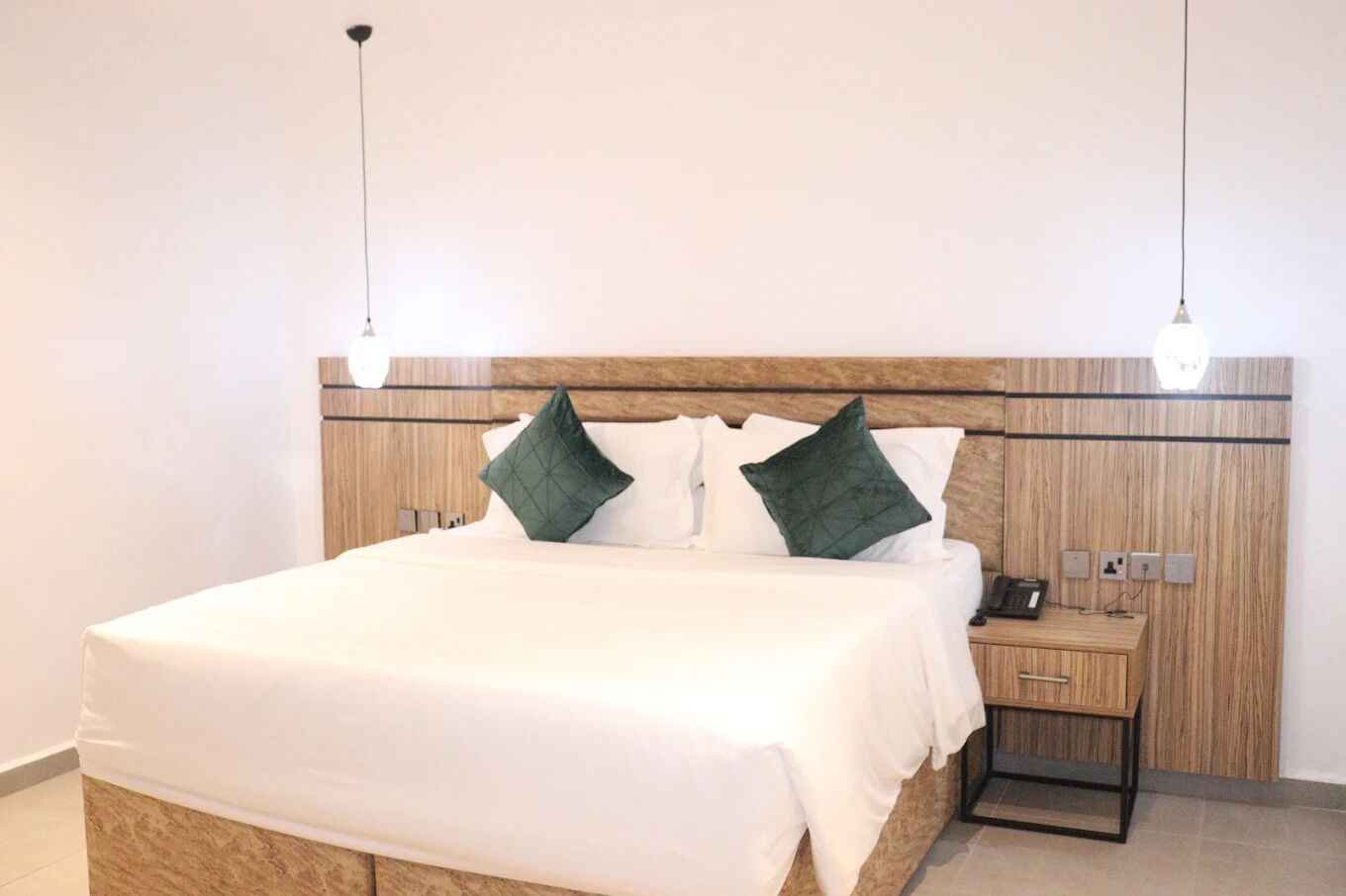 A neatly made bed with white linens and two green pillows, a wooden headboard, a bedside table with a phone, and two pendant lights.