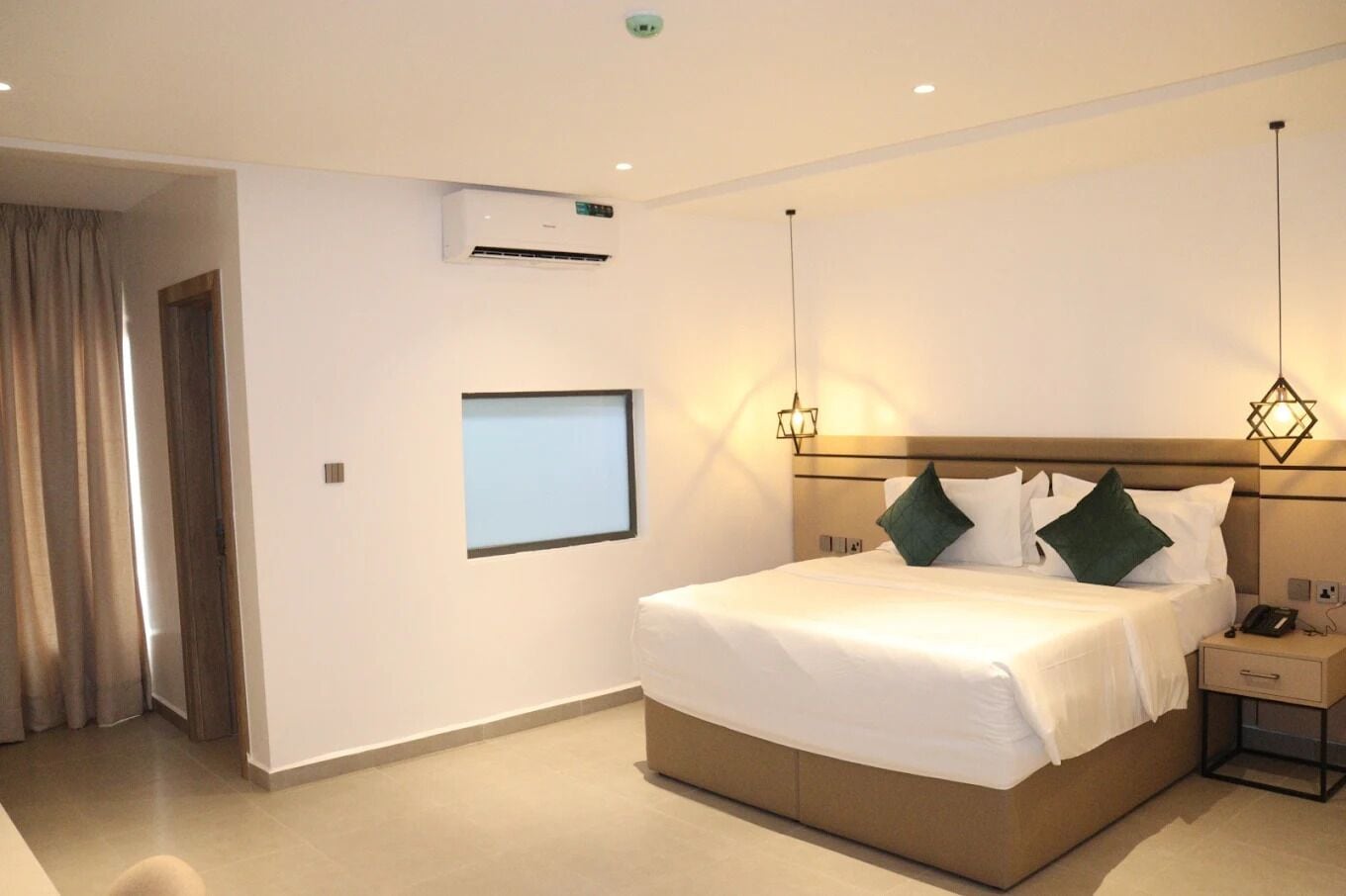 A hotel room with a large bed, bedside tables, a wall-mounted air conditioner, and a window with a view.