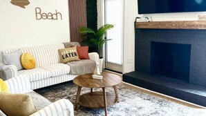 Een woonkamer met een gestreept bankje, een ronde salontafel, een open haard en een aan de muur bevestigde televisie waarop 'NETFLIX' wordt weergegeven.