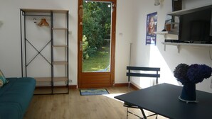 A room with a wooden door leading to a garden, a teal sofa, a small table with a vase, and a TV on a shelf. - "Chestnut, between land and sea (Plouézec)