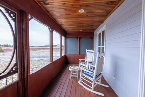 Een houten veranda met een schommelstoel en een kleine tafel, grote ramen en uitzicht over een veld.