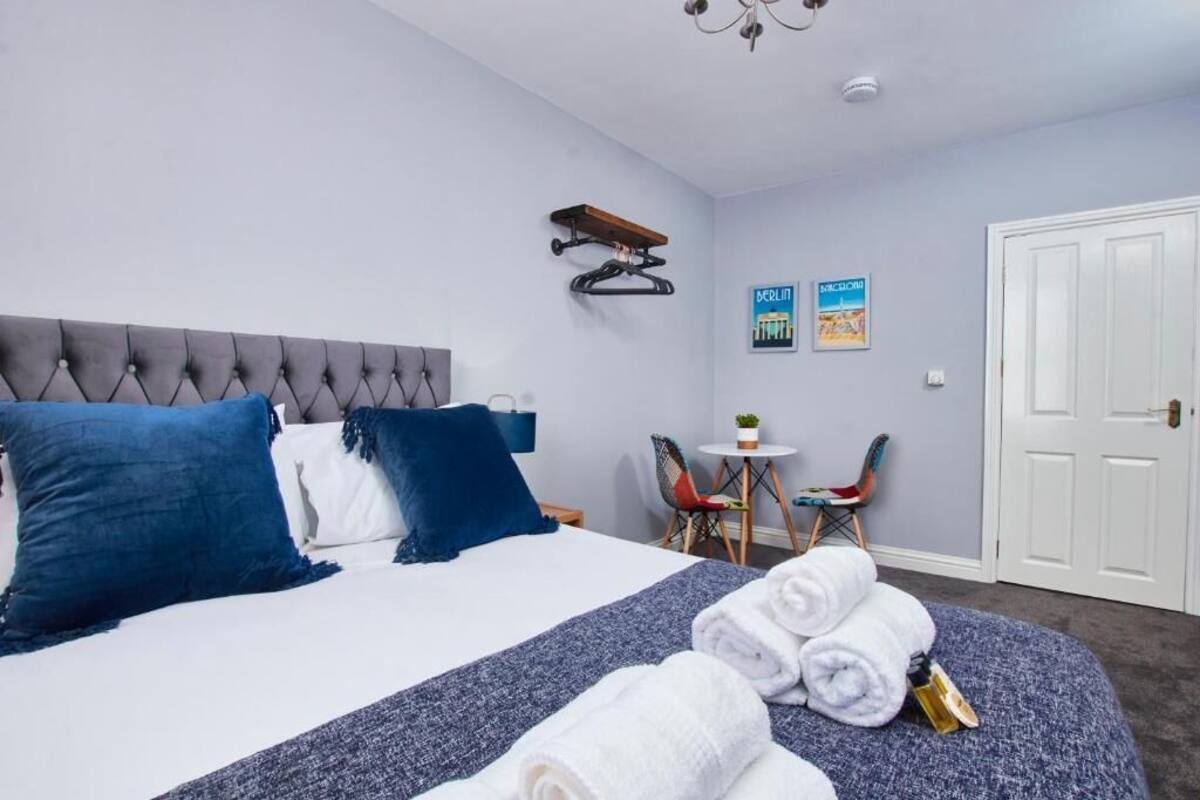 A neatly made bed with blue and white pillows, a small table with a plant, and a chair in a room with light blue walls and a white door.