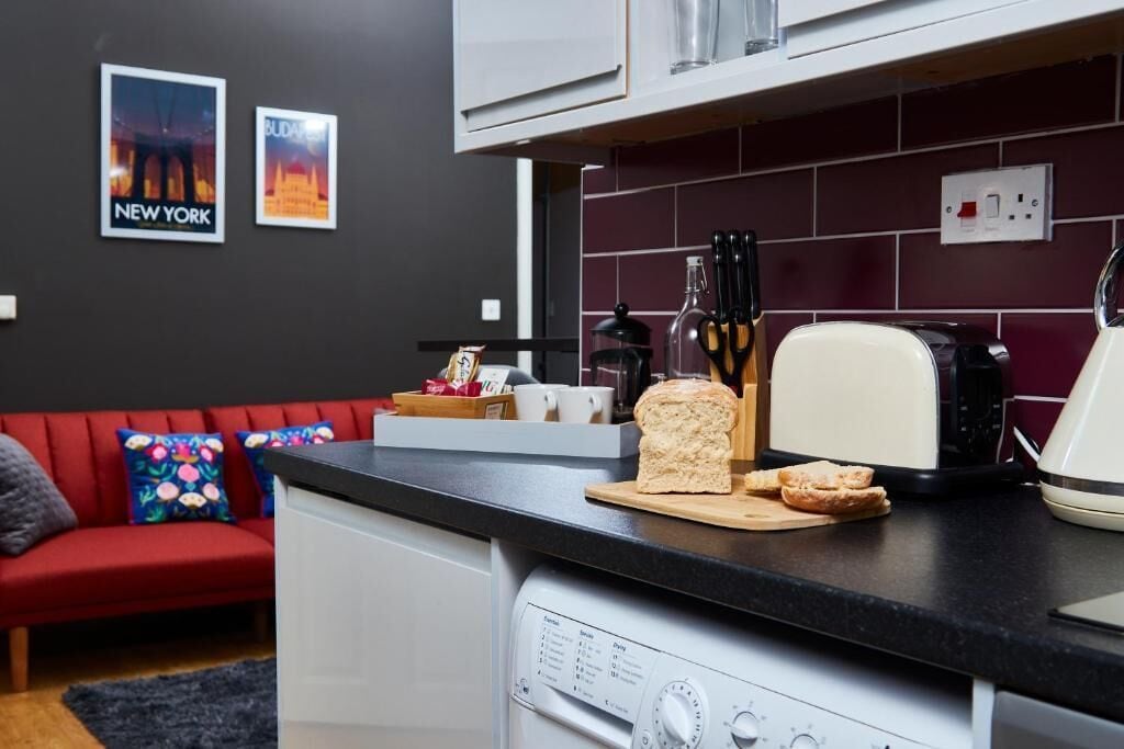 A compact kitchen with a toaster, a coffee maker, and a bread loaf on the counter. A red sofa and colorful pillows are visible in the background.