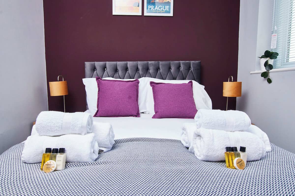 A neatly made bed with white and purple linens, two bedside lamps, and toiletries on the bed.