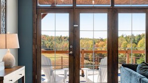 A room with large sliding glass doors leading to an outdoor deck with white chairs and a view of autumn foliage. - Calaboose Retreat- Hot Tub, Outdoor Pool, Pool Table, FirePit, Fireplace (Campton)