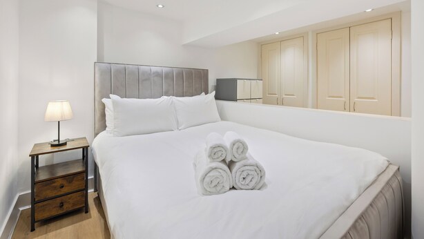 Cama bem arrumada com toalhas enroladas, mesa de cabeceira com abajur e guarda-roupa com portas de correr.