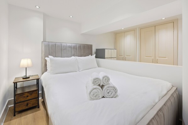 A neatly made bed with rolled towels, a bedside table with a lamp, and a wardrobe with sliding doors. - Spacious Fitzrovia 2-BR, Quiet & WiFi (London)