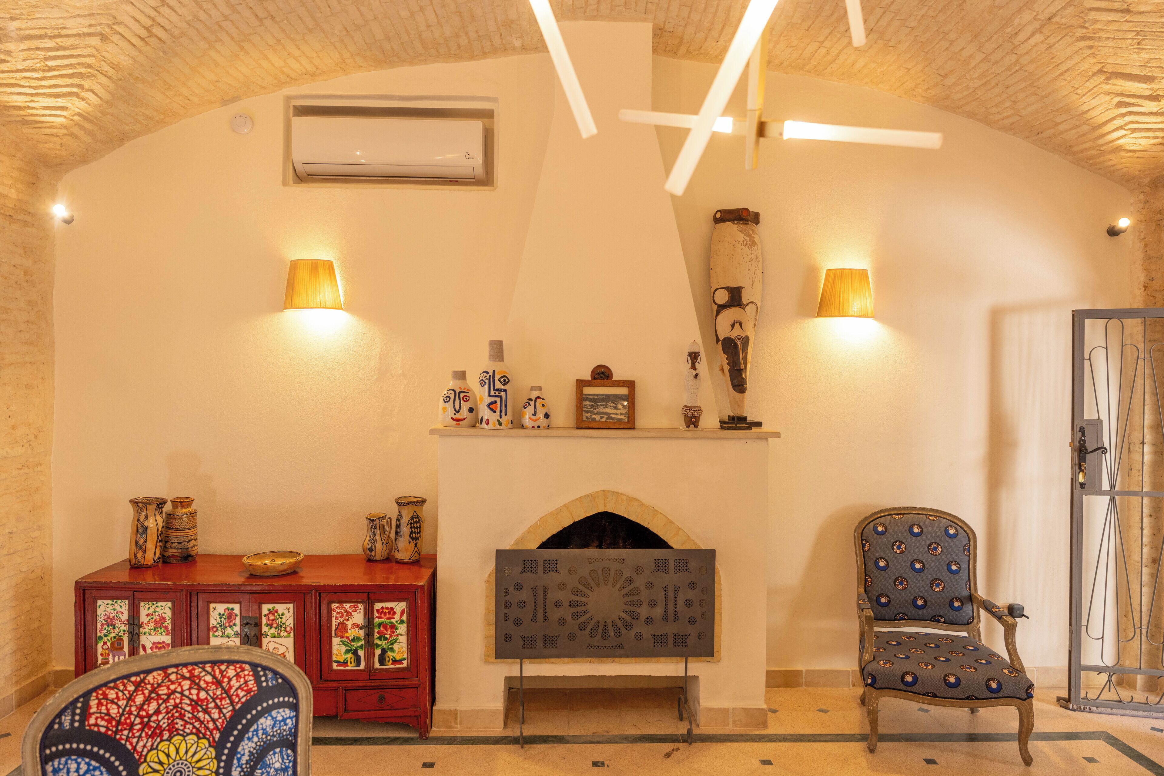 Une pièce avec une cheminée, deux lampes fixées au mur, un meuble décoratif orné de vases et un fauteuil à motifs.