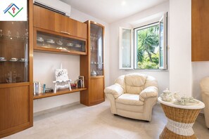 Ein Wohnzimmer mit einem beigefarbenen Sessel, einer Vitrine mit Teeservice, einem Korbtisch mit Teeservice und einem Fenster mit Blick ins Grüne.