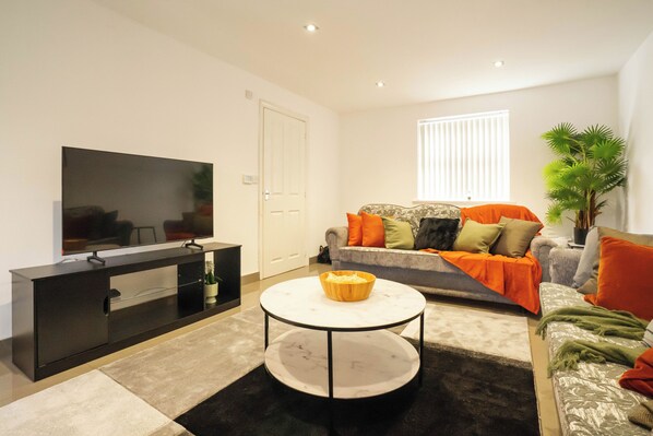 Una sala de estar moderna con un televisor de pantalla plana, un mueble de entretenimiento negro, una mesa de centro redonda, un sofá gris con cojines naranja y verde, y una planta en maceta.