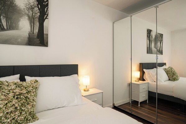 A modern bedroom with a large bed, a bedside table with a lamp, a nightstand, and a mirror.