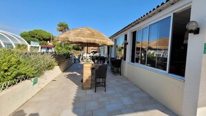 En patio med en stråtækt parasol, et bord og stole, et stort vindue og en grøn postkasse.