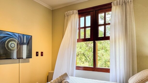 A hotel room with a bed, a television mounted on the wall, and a window with curtains. - Apê Bosque do Silêncio (Campos do Jordão)