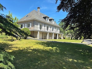 Une grande maison en pierre dotée d’un balcon, entourée d’une pelouse bien entretenue et d’arbres.