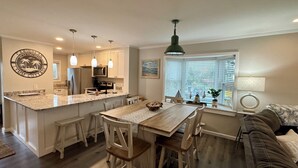 Une cuisine moderne dotée d’un îlot central, d’un coin repas avec des chaises en bois, d’un canapé et d’une grande fenêtre avec des stores.