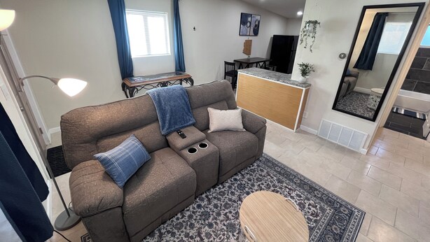 En moderne stue med sofa, sofabord og et lille rundt bord. Der er et spiseområde med bord og stole, et køkken med en køkkenø, og et badeværelse er synligt gennem en åben dør.