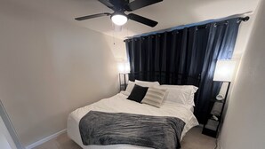 Et soveværelse med seng, ventilator, natbord, lampe og et vindue med gardiner.