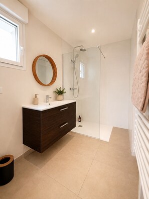 Une salle de bain moderne avec une douche à parois de verre, un meuble-lavabo en bois foncé, un miroir rond et un sol carrelé de couleur blanche.