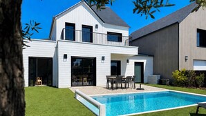 Ein modernes, zweistöckiges Haus mit Swimmingpool, einer Terrasse und Gartenmöbeln sowie ein großer Baum im Vordergrund.