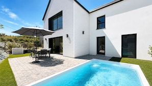 Una casa moderna con piscina, un patio con sillas y una sombrilla, y un césped bien cuidado.