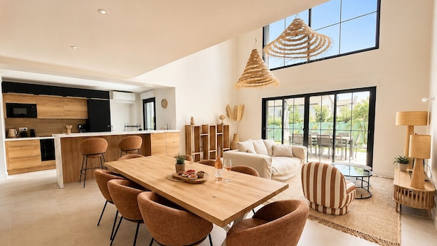 Una cocina moderna con una isla central, un comedor con mesa y sillas de madera, y una sala de estar con sofá y vistas al exterior.