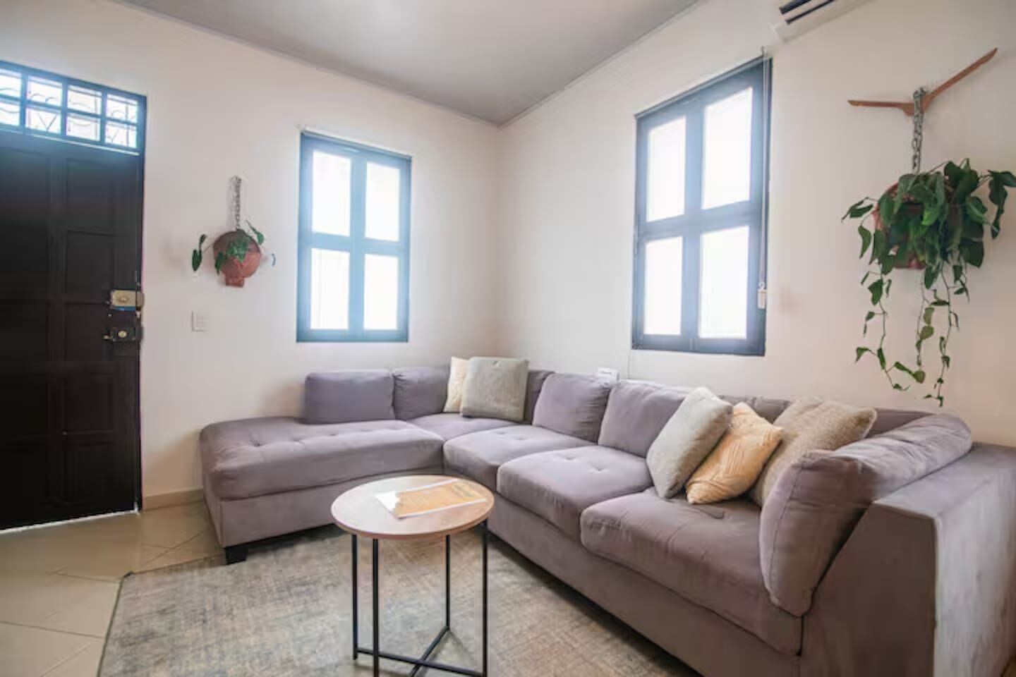 Ein Wohnzimmer mit einem grauen Ecksofa, einem kleinen runden Tisch und zwei Fenstern mit blauen Rahmen.