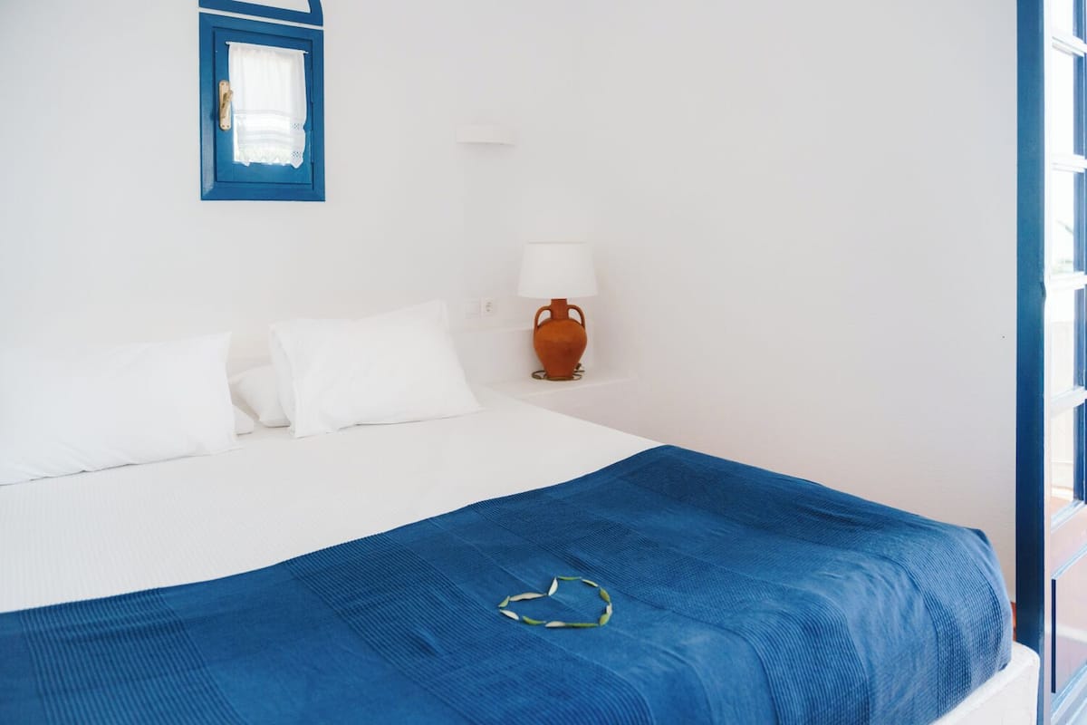 A bed with white bedding and a blue blanket, a white wall, a window with blue trim, and a brown vase with a white lamp on a nightstand.