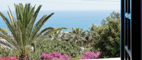 A balcony with a white table and chairs, overlooking a coastal landscape with palm trees and pink flowers.