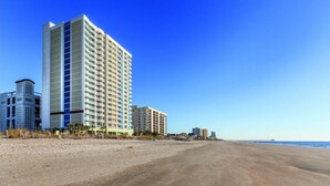 Un hôtel en bord de mer, avec plusieurs étages, une plage de sable en premier plan et un ciel bleu dégagé.