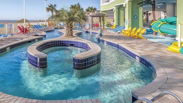 Un espace piscine avec des chaises longues bleues et jaunes, un palmier et un toboggan aquatique.
